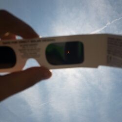 A hand holding up paper solar eclipse glasses against a bright blue sky, revealing a tiny orange crescent sun visible through the dark filter lens.